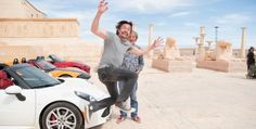 a man and woman jumping in front of a white sports car with statues behind them