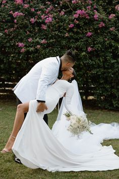 a bride and groom kissing in front of pink flowers at their wedding day, with one holding the other's leg