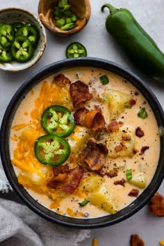 a black bowl filled with cheese and bacon soup next to green peppers, jalapenos