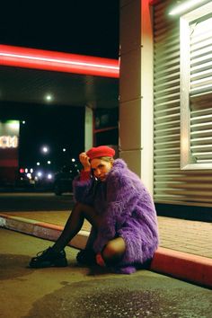 a person sitting on the ground in front of a building at night wearing a purple furry coat