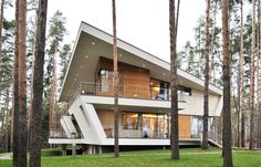 a modern house in the woods surrounded by tall pine trees and green grass, with stairs leading up to the upper level