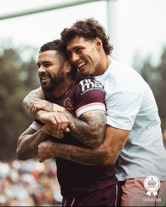 two men are hugging each other in the middle of a soccer game, one is wearing a white t - shirt and the other has tattoos on his arms