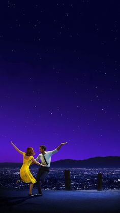 a man and woman dancing on top of a roof at night with the city lights in the background