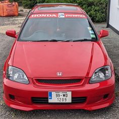 the front end of a red honda civic car parked on gravel with trees in the background