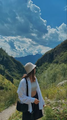 a woman in a hat and dress is standing on a path with mountains behind her