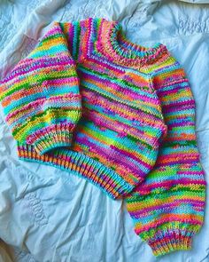 a colorful knitted sweater laying on top of a white bed next to a pillow