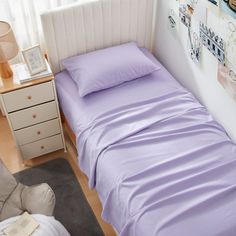 a small bed with purple sheets and pillows in a room next to a chair, lamp and pictures on the wall