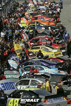 many cars are lined up in a row on the track, with people standing around them