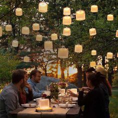 a group of people sitting around a table with candles hanging from the trees above them