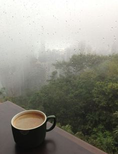 a cup of coffee sitting on top of a table next to a window covered in rain