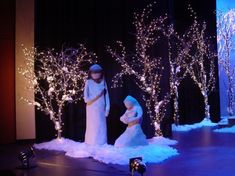 two women dressed in white dress standing next to each other on stage with trees and lights behind them