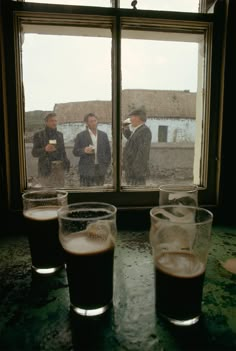 three men standing in front of a window with beer glasses on the table next to them