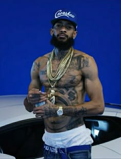 a man with tattoos standing in front of a white car wearing a hat and chain