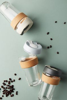 two glass cups with lids are next to some coffee beans on a green surface, one is empty and the other has a cork lid