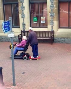 an elderly man pushing a woman in a motorized scooter on the sidewalk with a sign that says, relationship goals find someone lazy