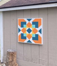 an orange and blue quilt hanging on the side of a building next to a tree stump