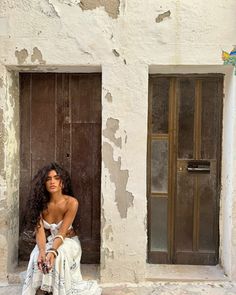 a woman sitting on the ground in front of a building with peeling paint and wooden doors