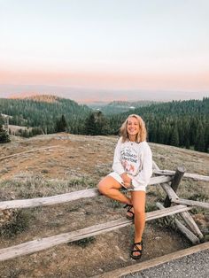 a woman sitting on top of a wooden bench in front of a field and forest