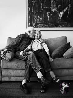 an older couple sitting on a couch in front of a large painting and stuffed animal