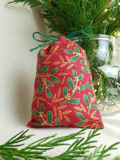 a red bag with green leaves on it sitting next to a pine branch and glass vase