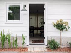 an open door to a white house with plants in the foreground and a table on the other side