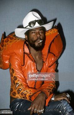 a man in an orange jacket and white cowboy hat sitting on a chair with his hands behind his head