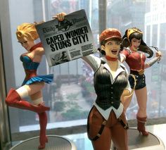 three figurines are posed in front of a glass display case, one holding a paper sign that says caper wonder stands city