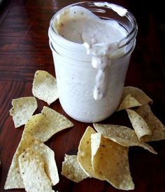 a glass jar filled with white dip surrounded by tortilla chips on a wooden table