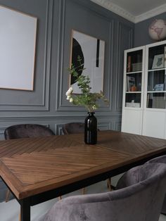 a wooden table topped with a vase filled with flowers next to two gray velvet chairs