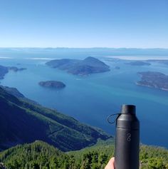 a person holding a water bottle in their hand overlooking the ocean and mountains with blue sky