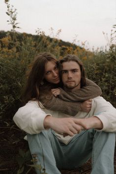 a man and woman sitting on the ground in front of some flowers with their arms around each other