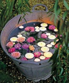 candles floating in a bucket filled with water surrounded by pink and red flowers on the ground