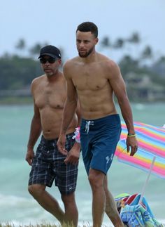 two shirtless men walking on the beach with an umbrella behind them and one man in plaid shorts