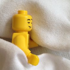 a yellow lego man laying on top of a white blanket with his head resting on the pillow