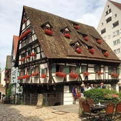 an old house with flowers on the roof and people walking around it in front of some buildings