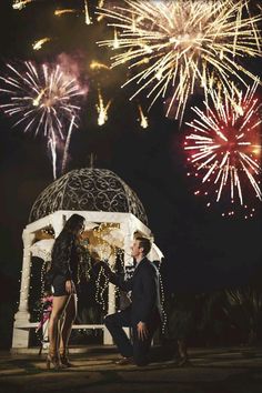 a man and woman standing next to each other with fireworks in the sky behind them