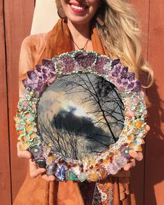 a woman holding up a mirror with trees in the middle and clouds on it,