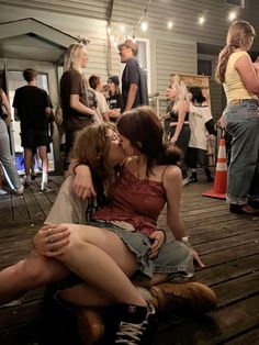 two women sitting on the ground kissing while others stand in the background and people are standing around