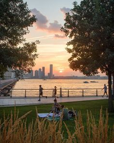 some people are sitting on the grass by the water and watching the sun go down