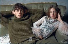 a man and woman laying on a couch with their arms behind each other, both wearing sweaters