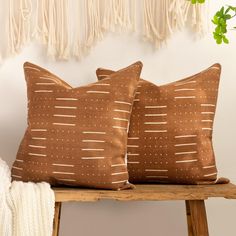 two brown pillows sitting on top of a wooden bench next to a wall hanging with tassels