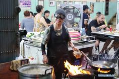 a woman cooking food over an open fire in a kitchen with many other people around