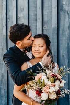 a bride and groom hugging each other in front of a wooden wall with flowers on it