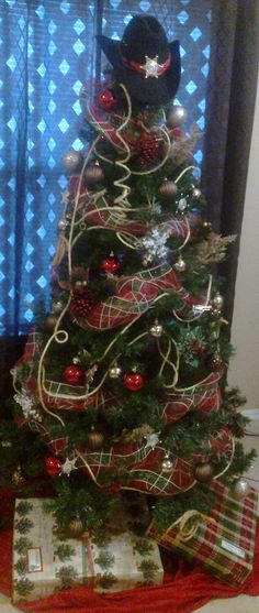 a christmas tree with presents under it and a cowboy hat on the top, in front of a window