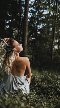 a woman sitting in the middle of a forest looking up into the sky with her hair blowing back