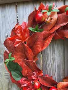 a wreath made out of red leaves and apples hanging on a wooden fence with green leaves