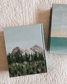 two books sitting on top of a bed next to each other with mountains in the background