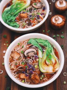 two bowls filled with noodles and vegetables on top of a wooden table next to mushrooms