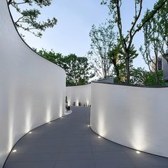 an outdoor walkway with white walls and lights on the sides, leading to trees in the distance