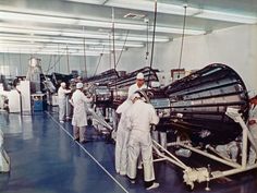 men in white suits working on an assembly line with large metal objects hanging from the ceiling